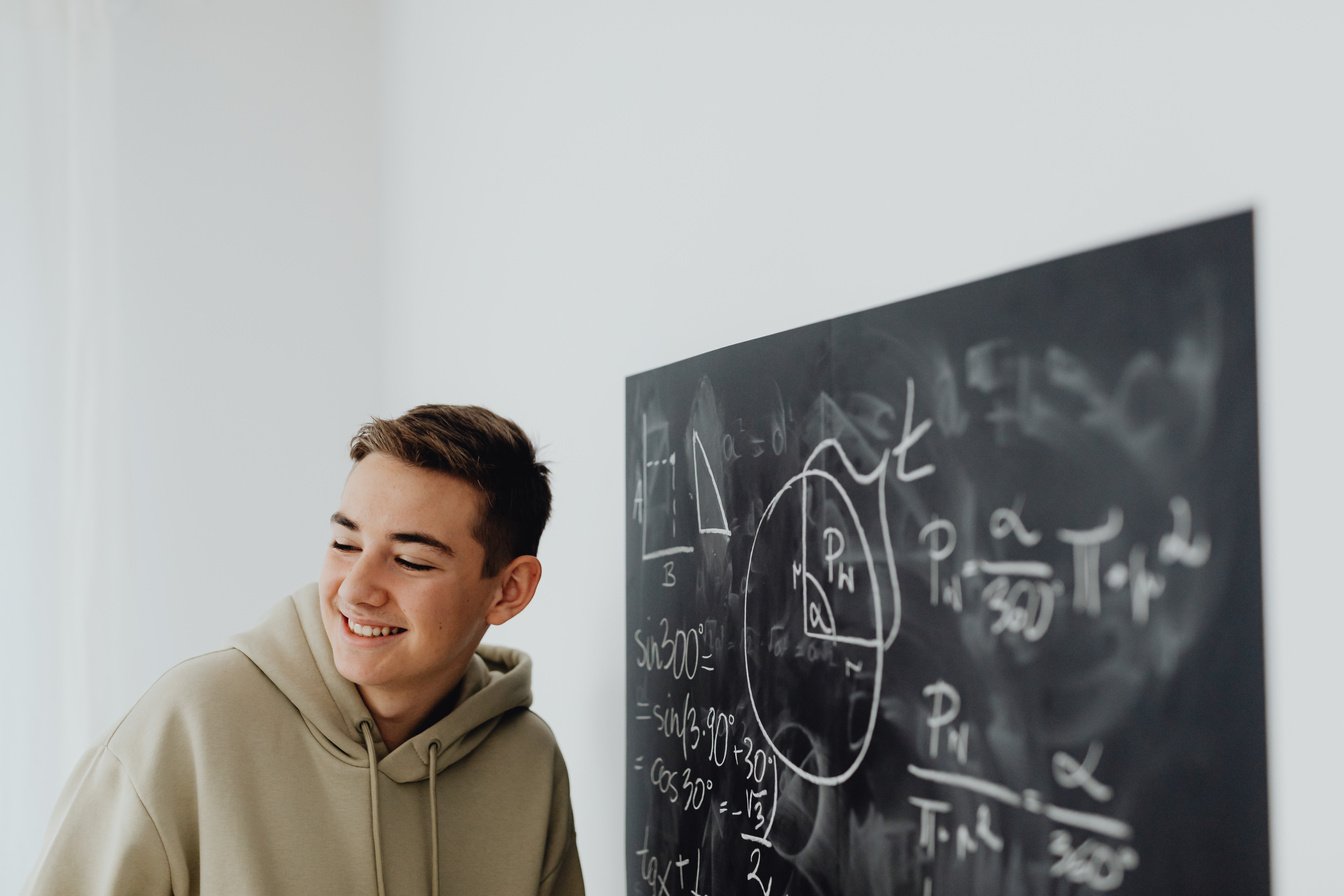 A Boy in Beige Hoodie Smiling Beside the Blackboard